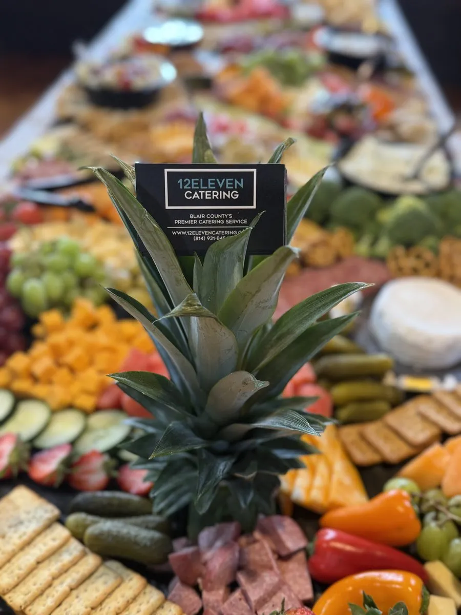 A 12Eleven Catering business card propped on a fresh pineapple centerpiece above a grazing table
