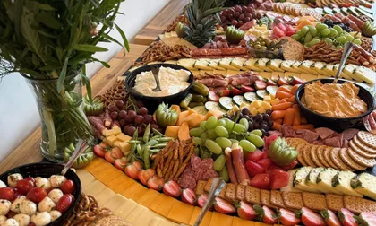 Grazing table set with cheeses, cured meats, dips, fresh fruit and crackers beside a floral arrangement